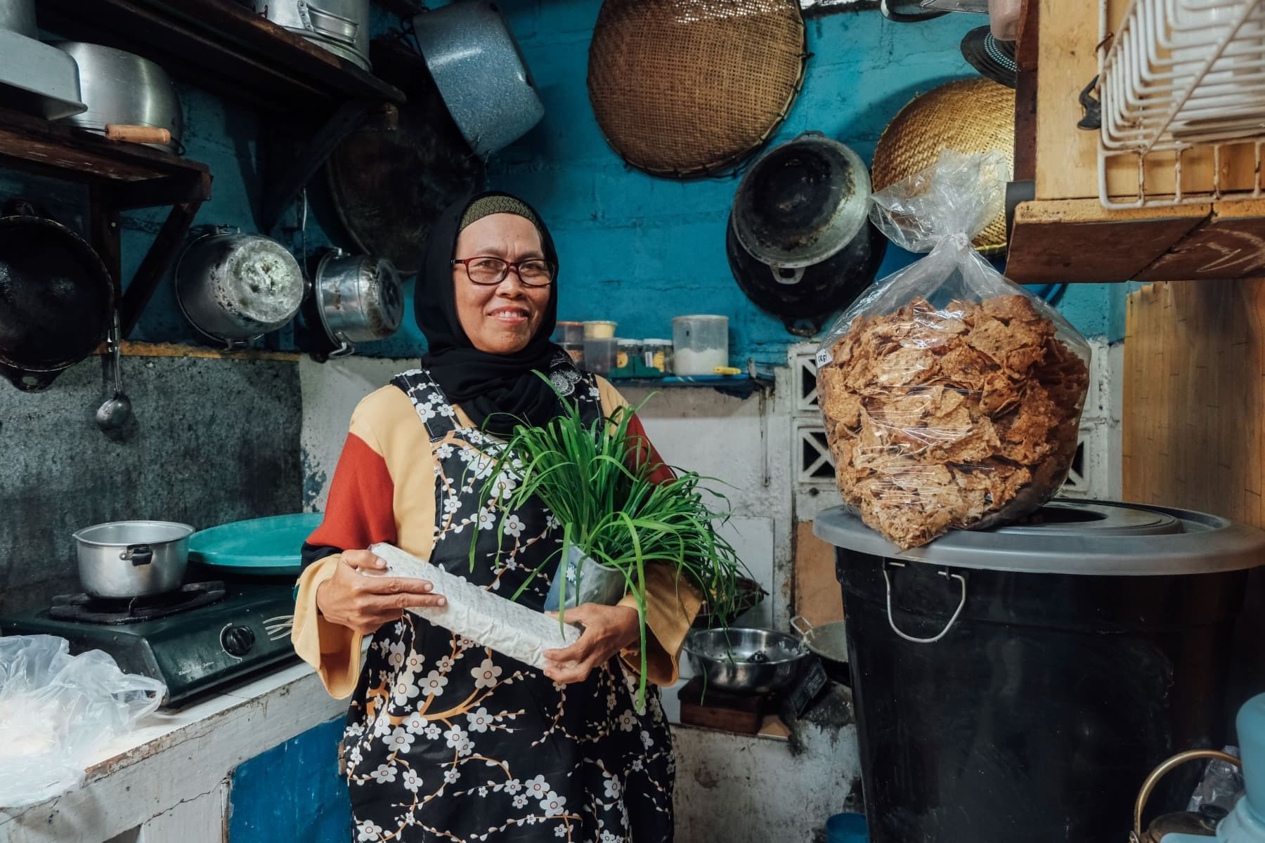 Foto Ibu Rustini tersenyum dengan membawa kucai dan tempe sebagai bahan baku Keripik Masshita.