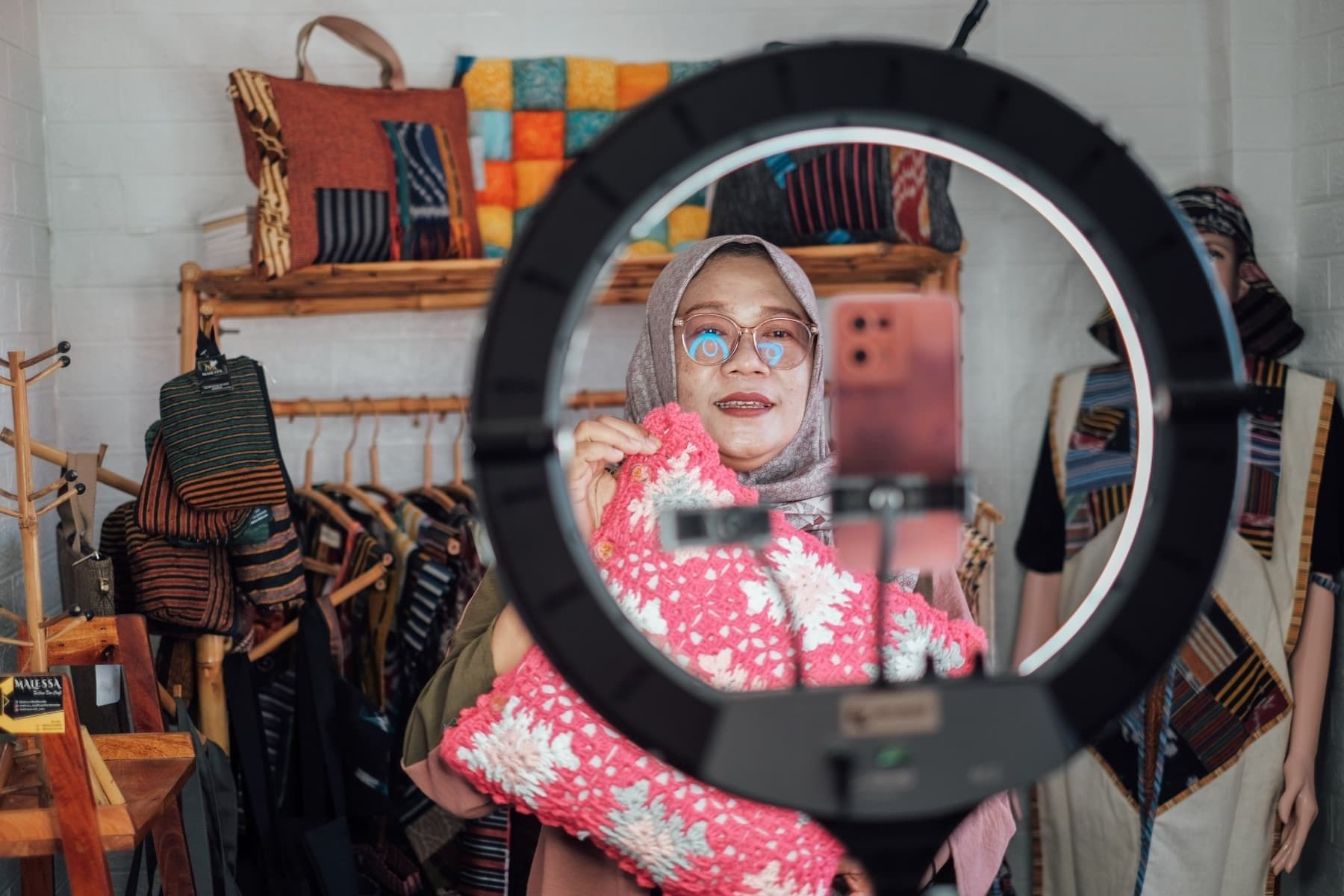 Foto Ibu Madu mempromosikan bantal rajut merah muda melalui live streaming di tokonya.